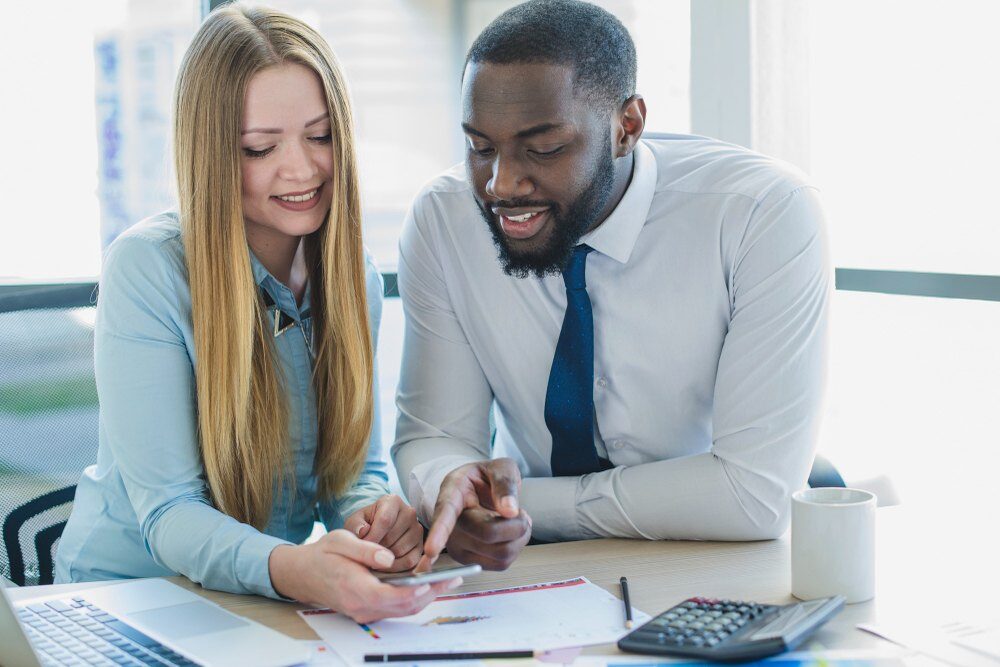 um homem e uma mulher apontando para um celular com notebook e calculadora ao lado